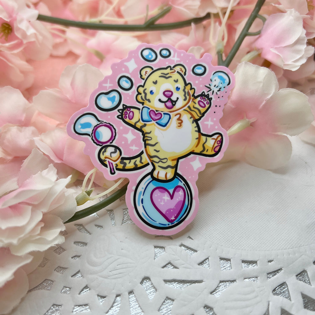 A sticker of a cute tiger balancing on a ball, bubble wand in tail, reaching up and poking the bubbles with his claws. Pink aesthetic overall.