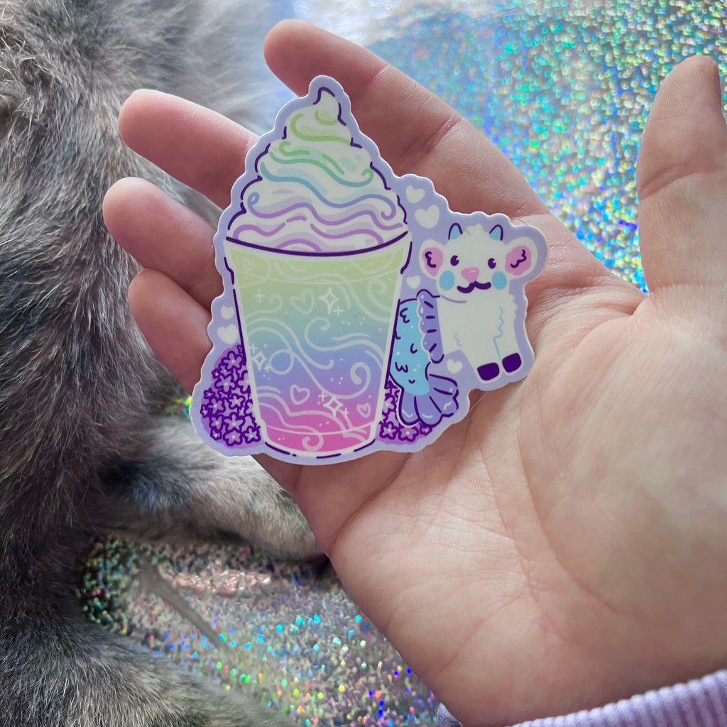 Hand holding a colorful sticker of a rainbow-themed drink and mermaid goat with a glittery background