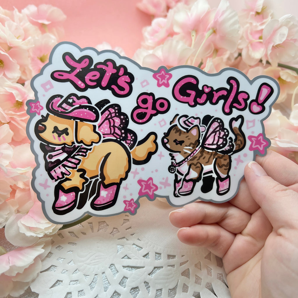 A bumper sticker of Two animals- one is a golden retreiver with a pink cowgirl hat, boots, and dark pink fairy wings. The second is a brown and white tabby cat with a white cowgirl haat, pink boots, and light pink butterfly wings. There is text that says "lets go girls!"