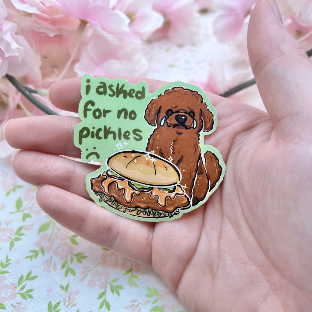 A sticker of a brown poodle dog sad and crying in front of a chicken sandwich that has pickles. Text says "i asked for no pickles :(" against a green background