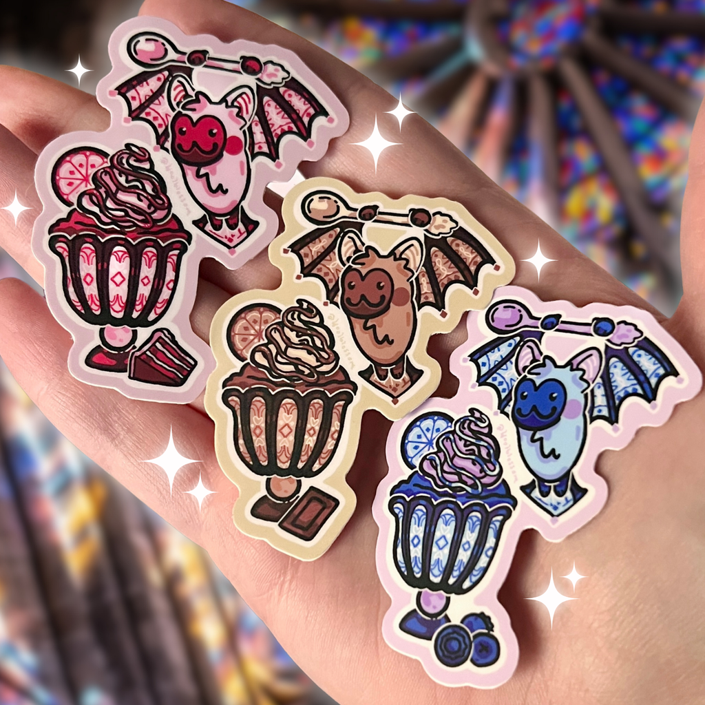 Hand holding 3 colorful bat-themed stickers that are stain glass and ice cream themed with a blurred background 