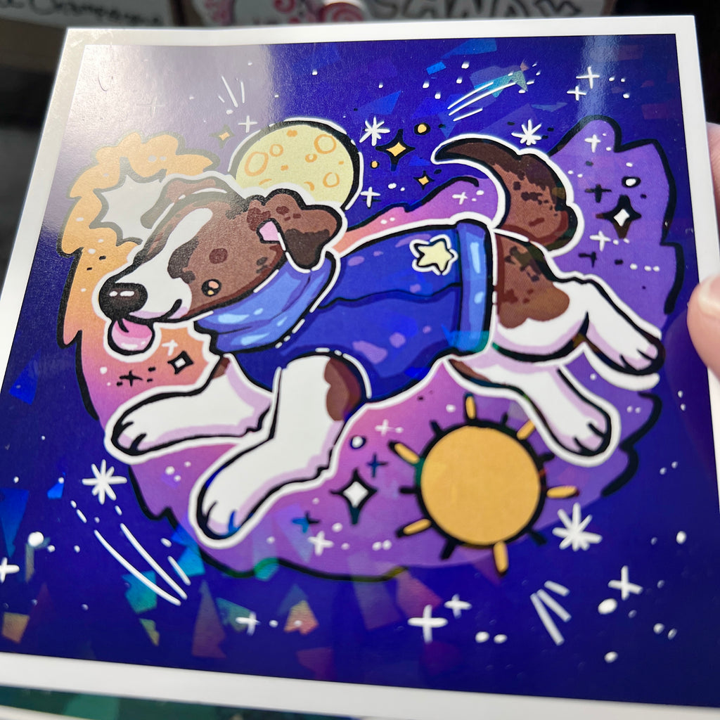 Print of a dog in a space-themed outfit on a glittery background held by hand
