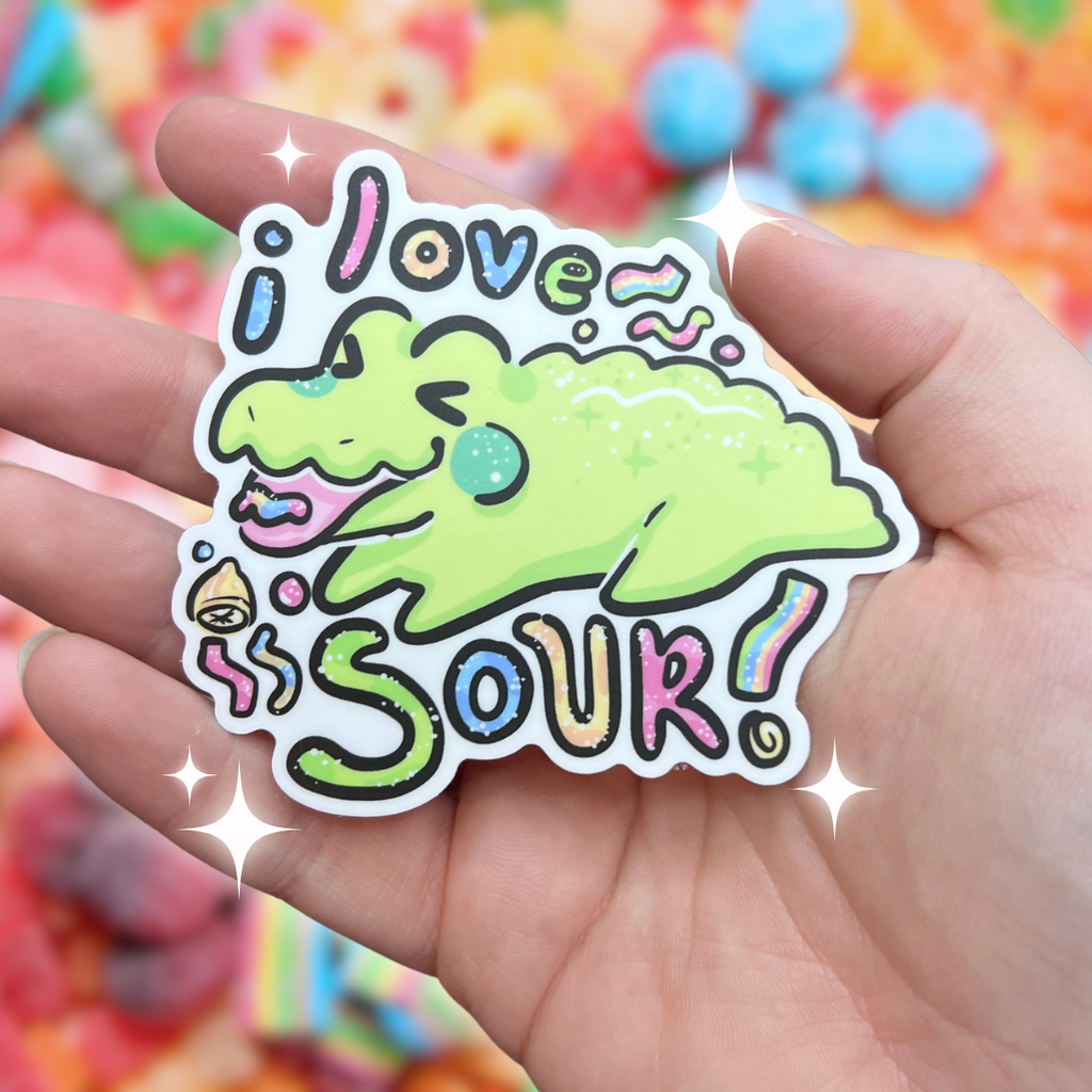 A sticker of a lime green, sparkly crocodile with a pile of sour candy in front of him, making a silly face, with text that reads "i love sour!" which is also sparkly like its candy