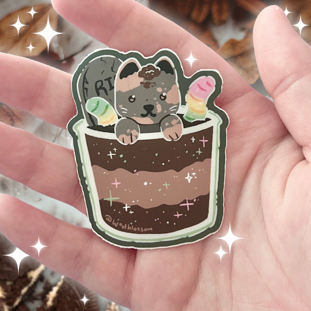 Sticker of a cute cat in a dirt cup with worms held in a hand with a blurred background.