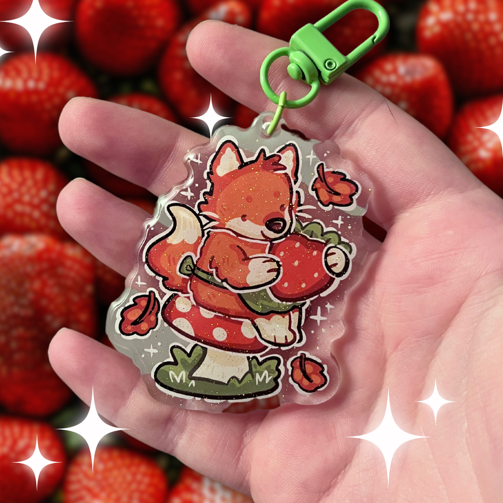 A keychain of a red fox with a little green apron sitting on a red mushroom, holding a strawberry with autumn leaves falling