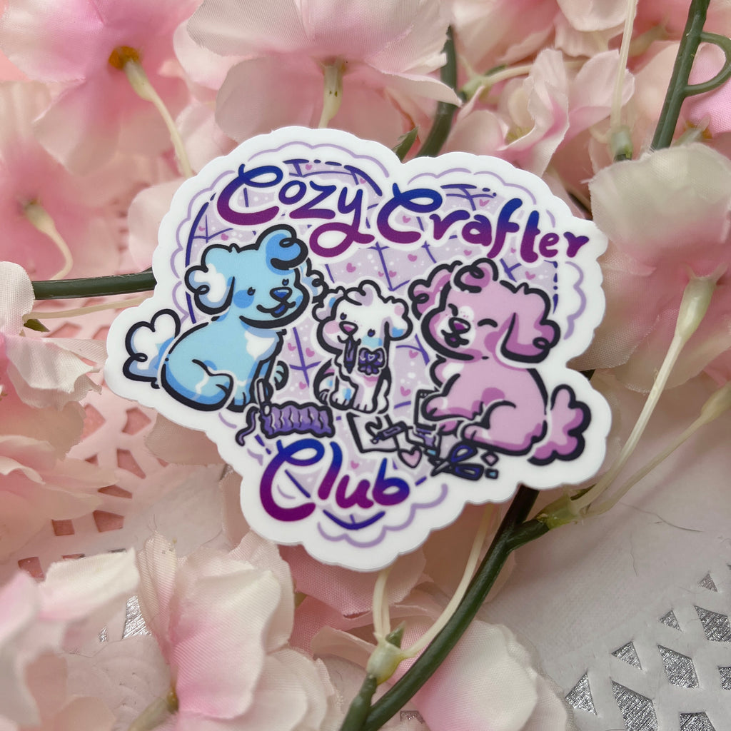 a sticker of three puppies who are knitting, doodling, and scrapbooking. One puppy is blue, one pink, and one white. There is text that says "cozy crafter club".