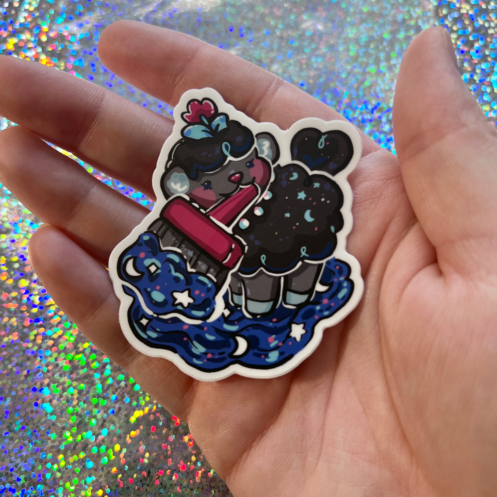 Hand holding a colorful sticker of a sheep holding a paintbrush on a glittery background