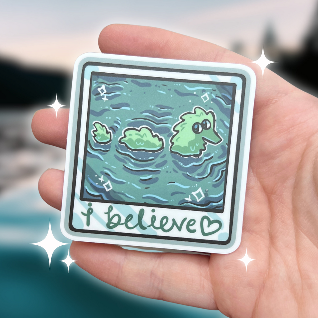 A sticker that looks like a camera photo, hand written text at the bottom that says "i believe <3", in the photo is lake water with a fuzzy worm lochness monster