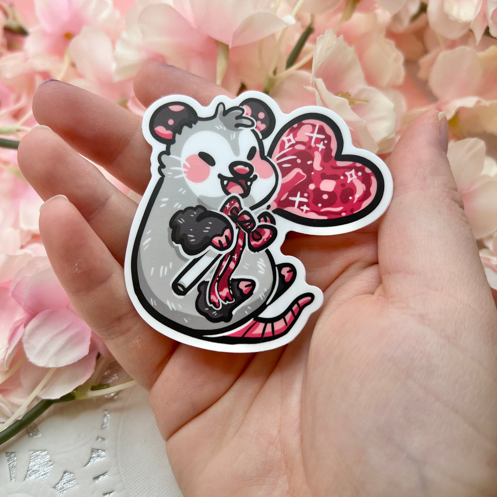 A sticker of an opossum happily holding a red heart lollipop