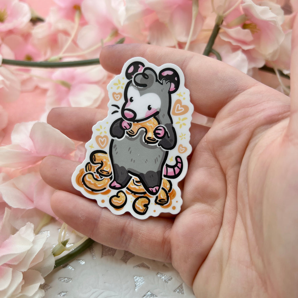 A sticker of an opossum standing in front of a pile of mac and cheese, holding one in its arms up to its mouth with hearts