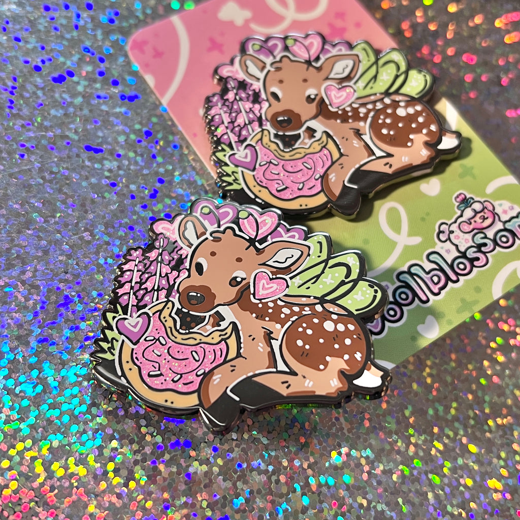 An enamel pin of a fairy deer eating a sugar cookie on a glittery background.