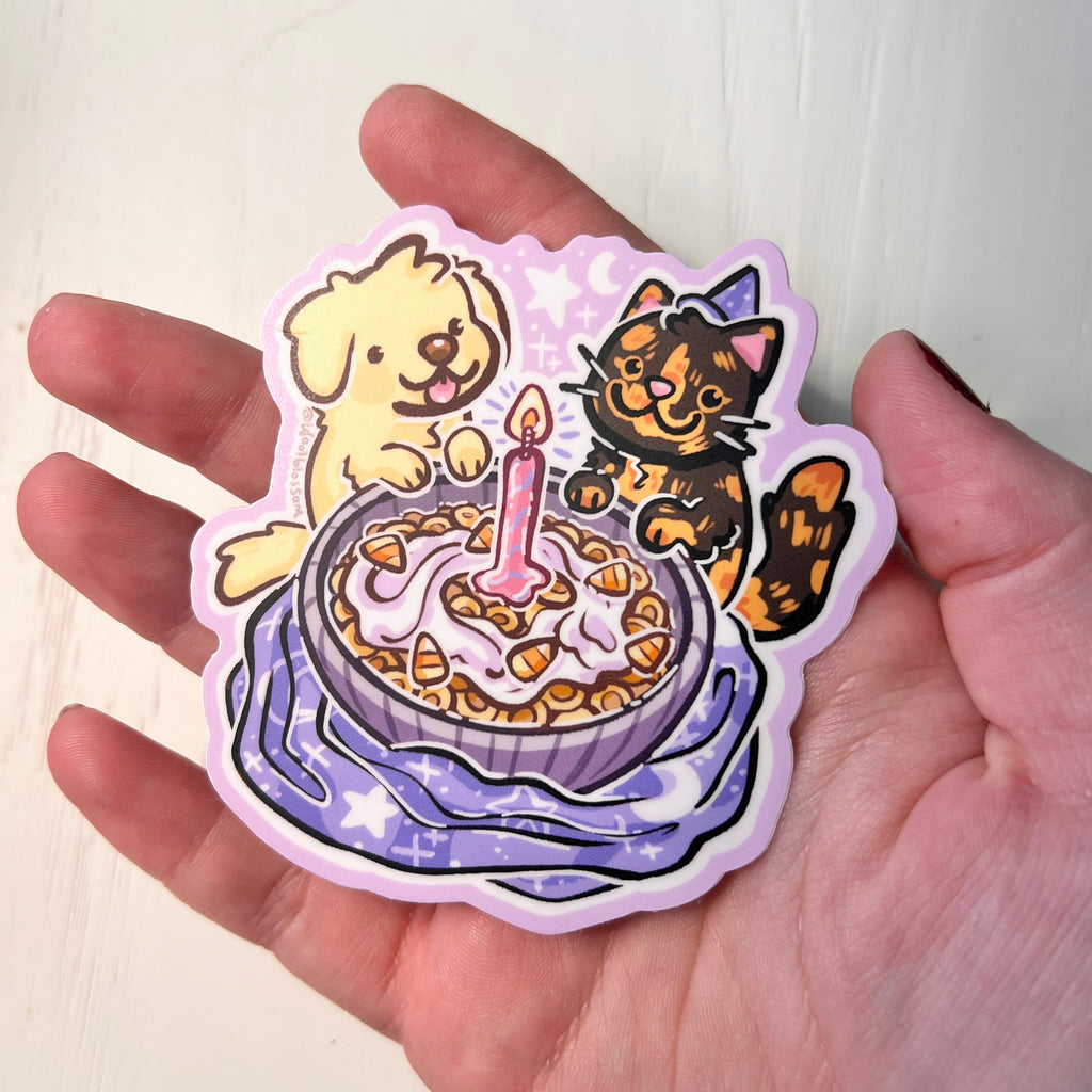 Sticker of a dog and cat by a birthday cereal with a candle held by a hand
