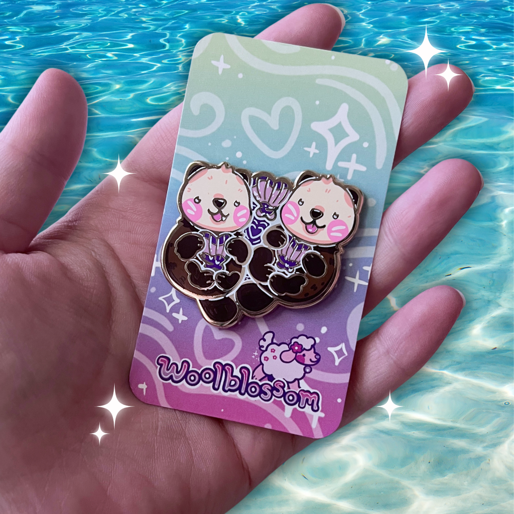 An enamel pin of two otters floating together holding heart shaped seashells
