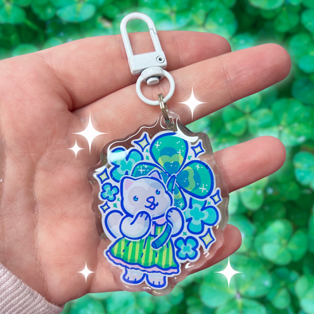 A keychain of a grey cat in a green stripe dress holding a four leaf clover