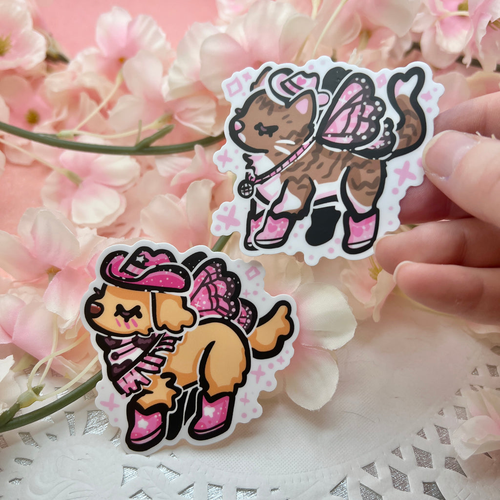 Two stickers- one is a golden retreiver with a pink cowgirl hat, boots, and dark pink fairy wings. The second is a brown and white tabby cat with a white cowgirl haat, pink boots, and light pink butterfly wings.