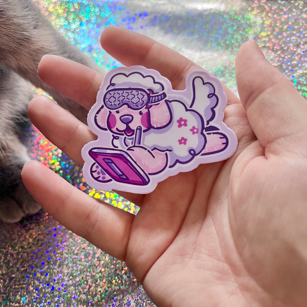 Hand holding a colorful sticker of a cartoon dog with sleep mask on drawing on an ipad, against a glittery background.
