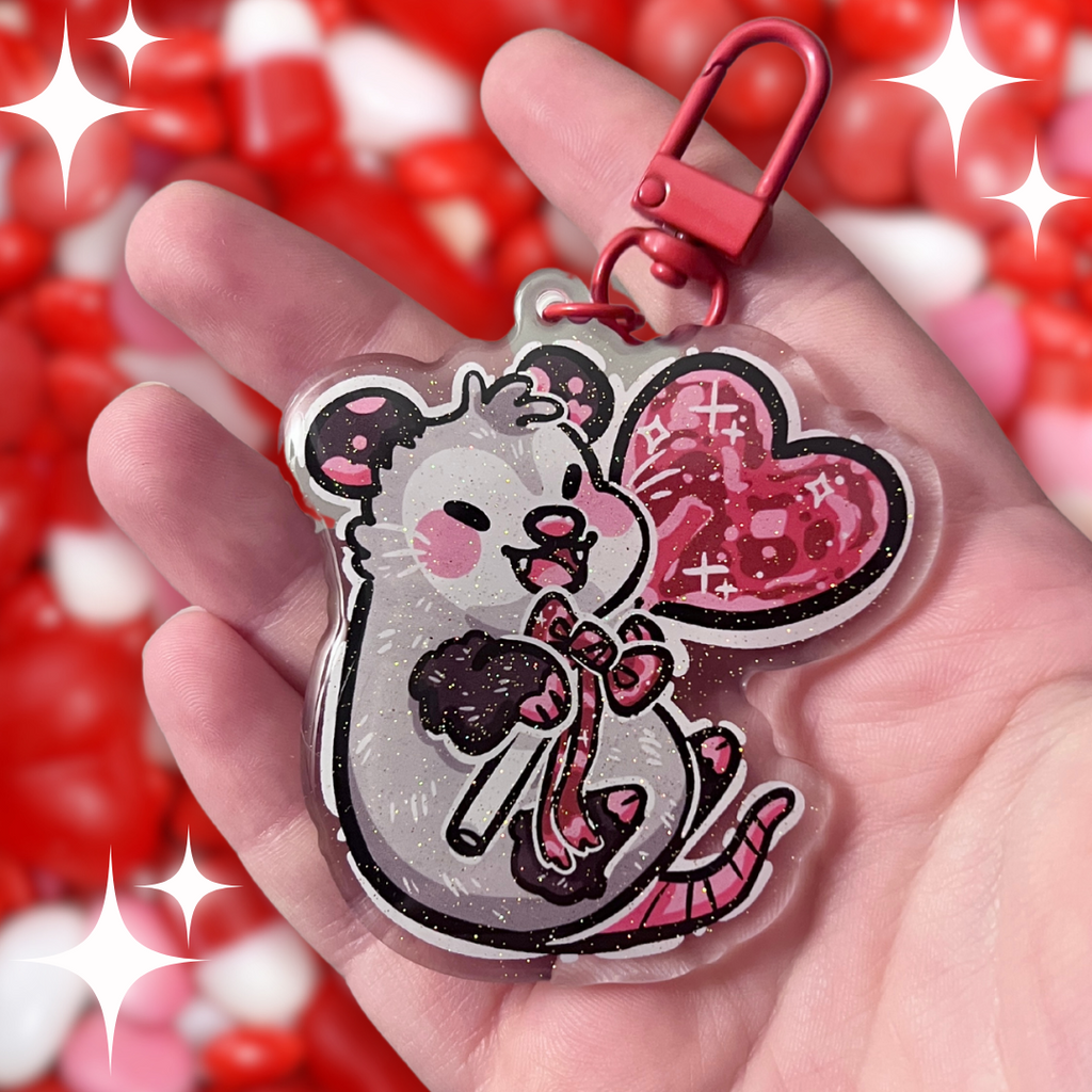 a keychain of an opossum happily holding a red heart lollipop