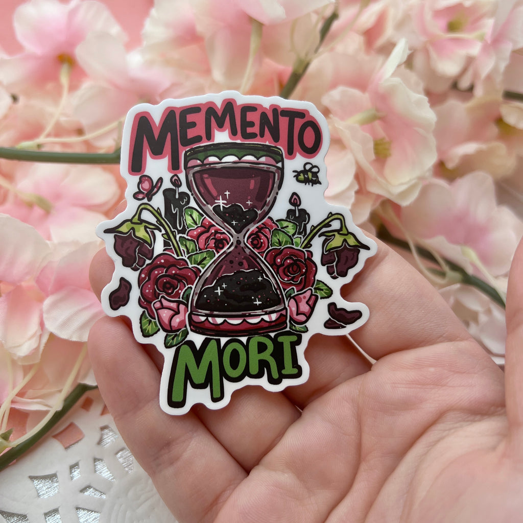 A sticker of a red and black hour glass surrounded by roses at various stages of their lifespan with text that says “memento mori”