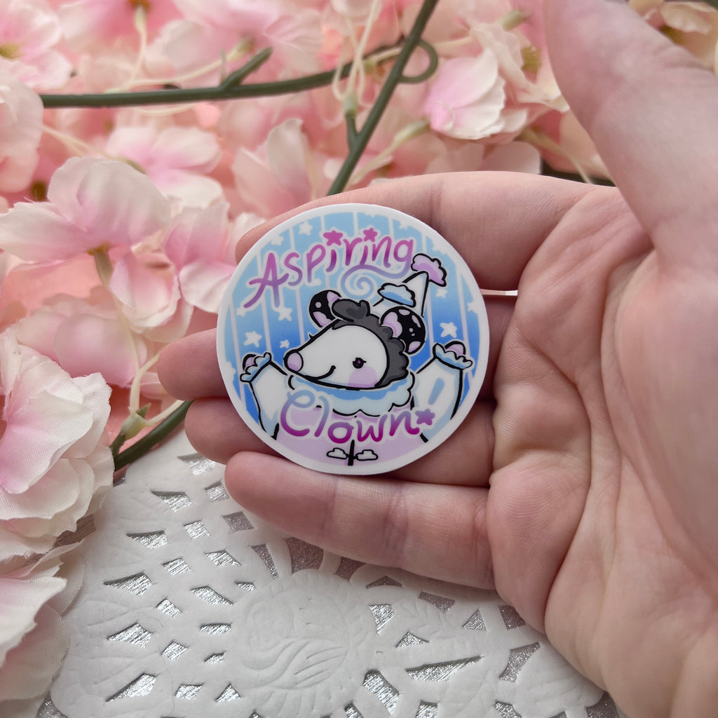 A sticker of a cloud themed opossum that looks happy with the text "aspiring clown"