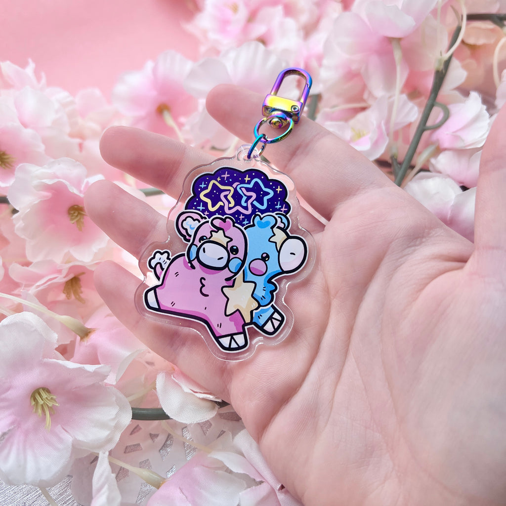 A keychain charm of a two headed calf, one is blue and one is pink, with a yellow star pattern connecting them and on each forehead. On top is a starry sky with three interlocking stars, one blue, one pink, and one yellow.