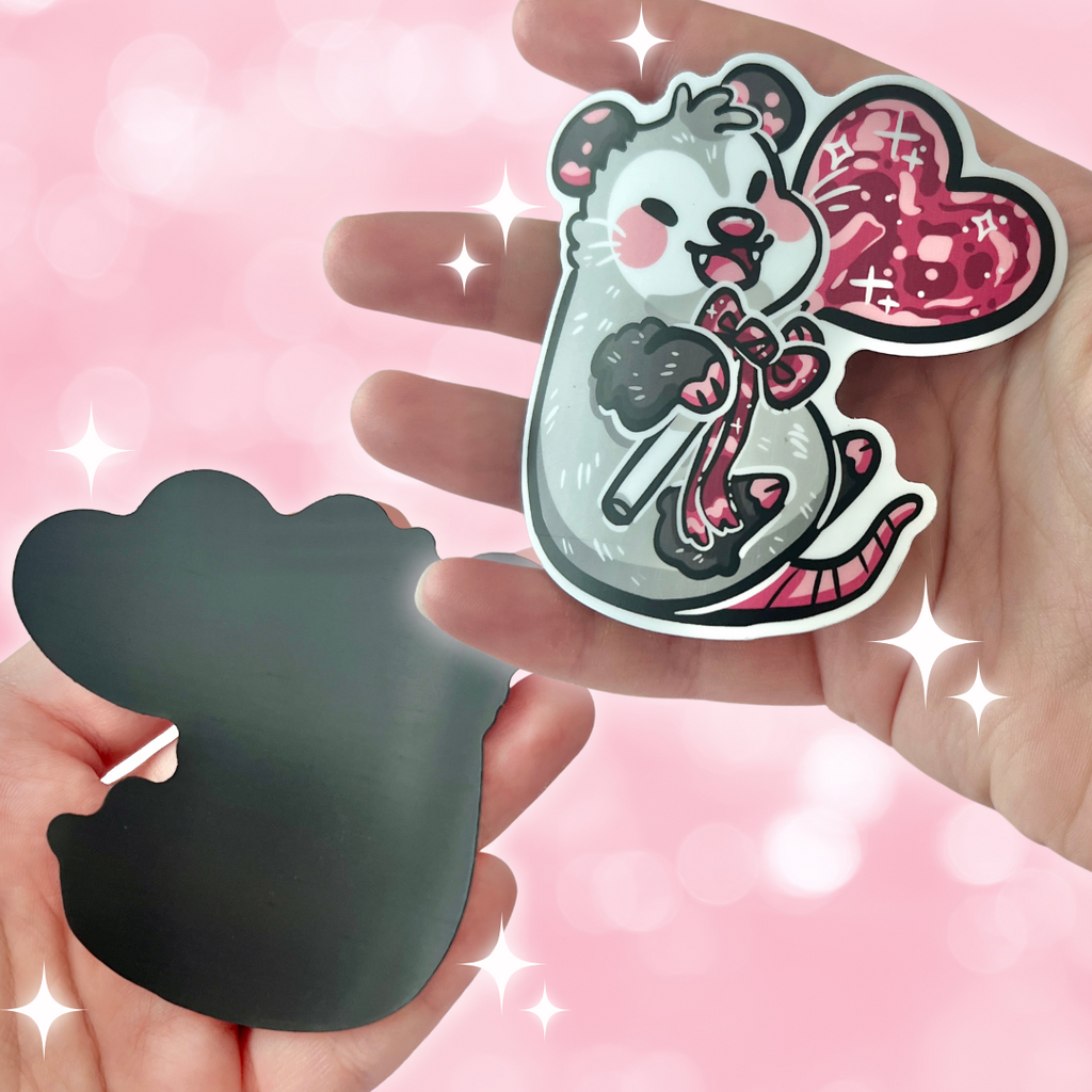 A magnet of an opossum happily holding a red heart lollipop