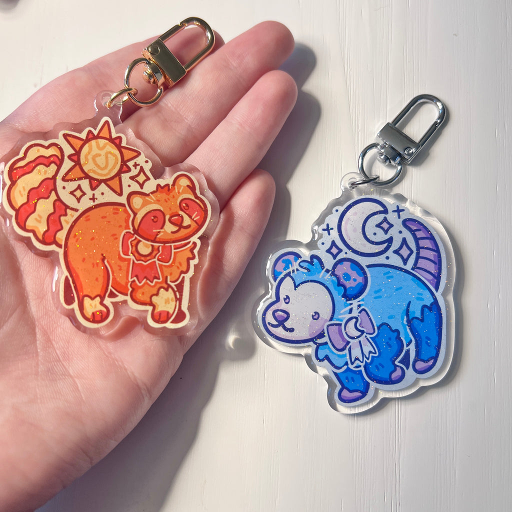 Two colorful keychains featuring cartoon sun raccoon and moon opossum held in a hand against a white background.