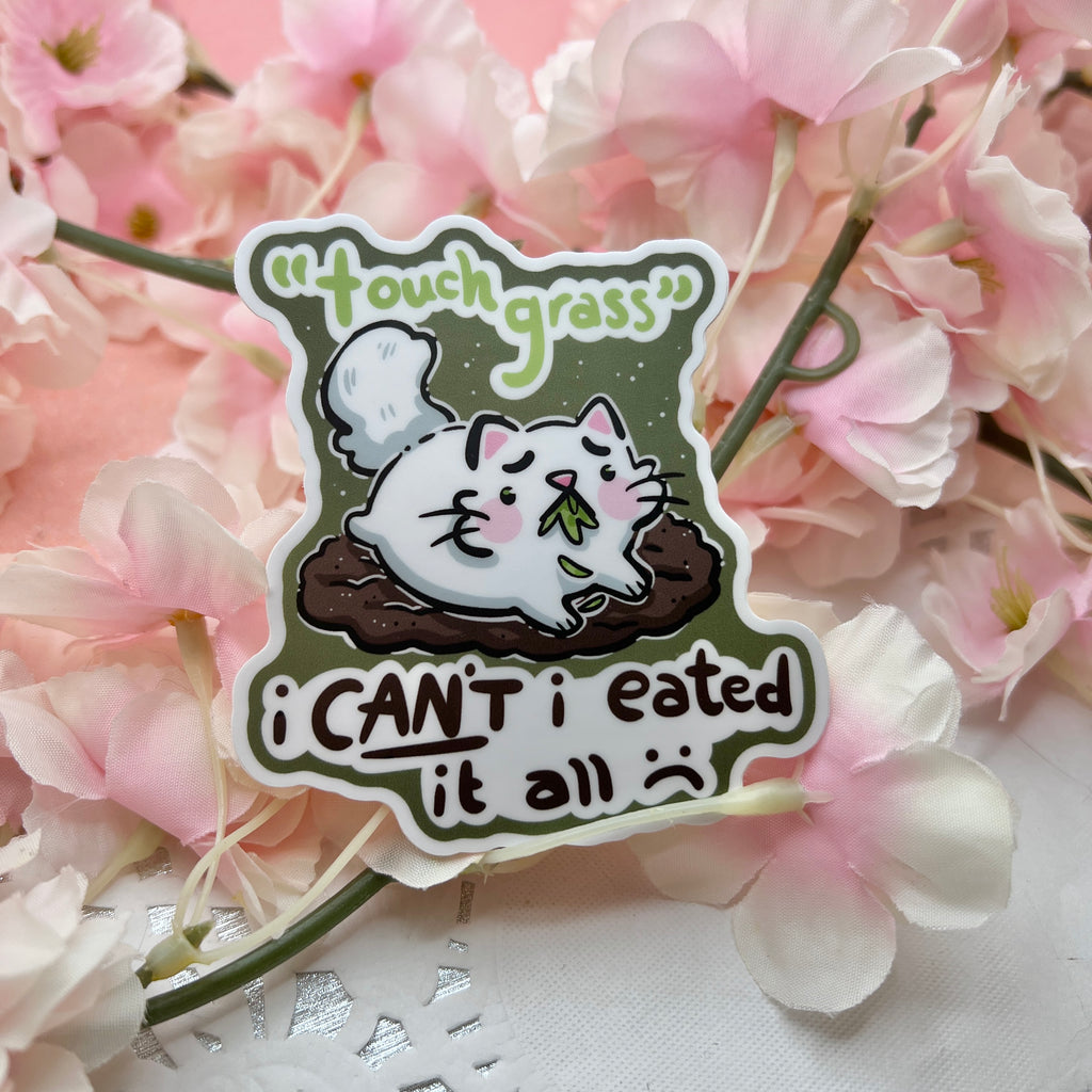 A sticker of a distressed cat laying on a bare patch of dirt, grass in her mouth with text that says "touch grass, i can't i eated it all :("