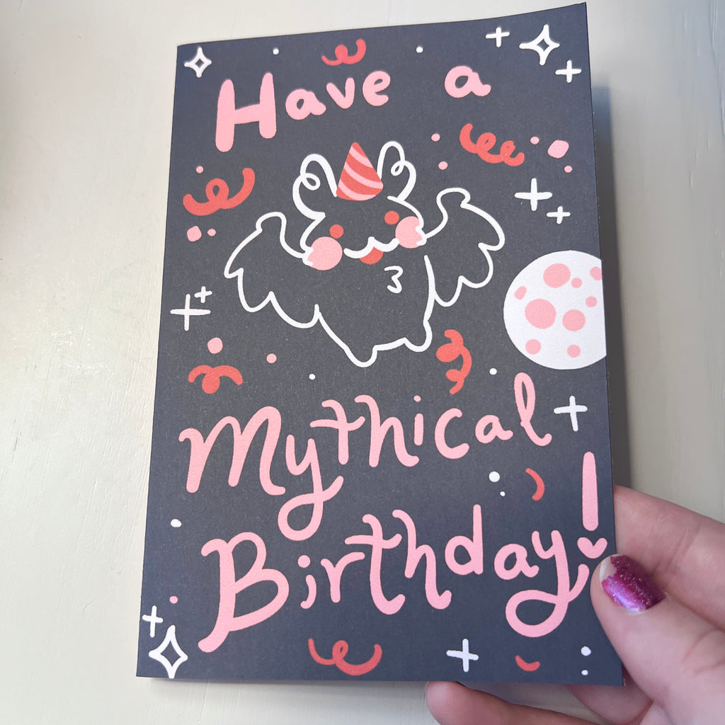 Hand holding a card with a mothman wearing a birthday hat and text 'Have a mythical birthday!' on a dark background.