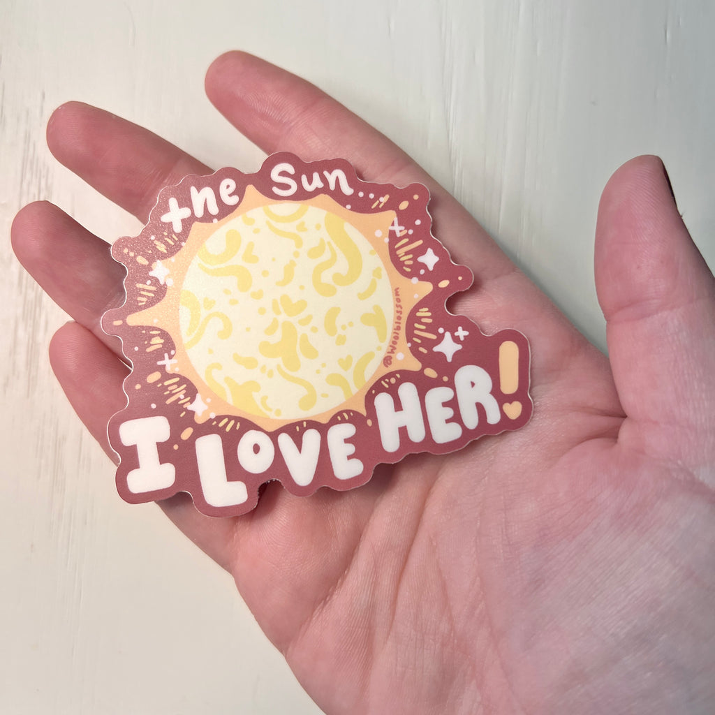 Sticker with sun design and text 'I Love Her' held in a hand against a white background