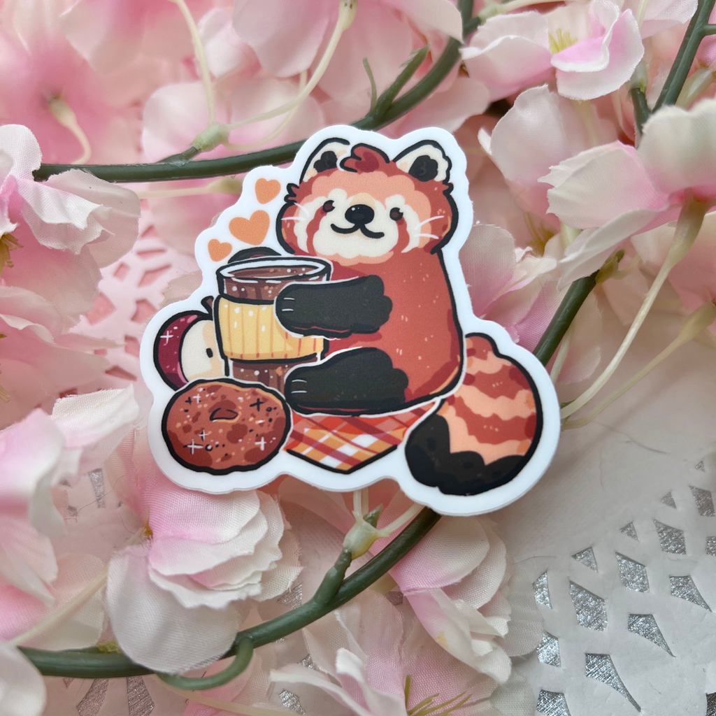 A sticker of a red panda holding a coffee drink with an apple, apple cider doughnut, on a warm toned plaid blanket