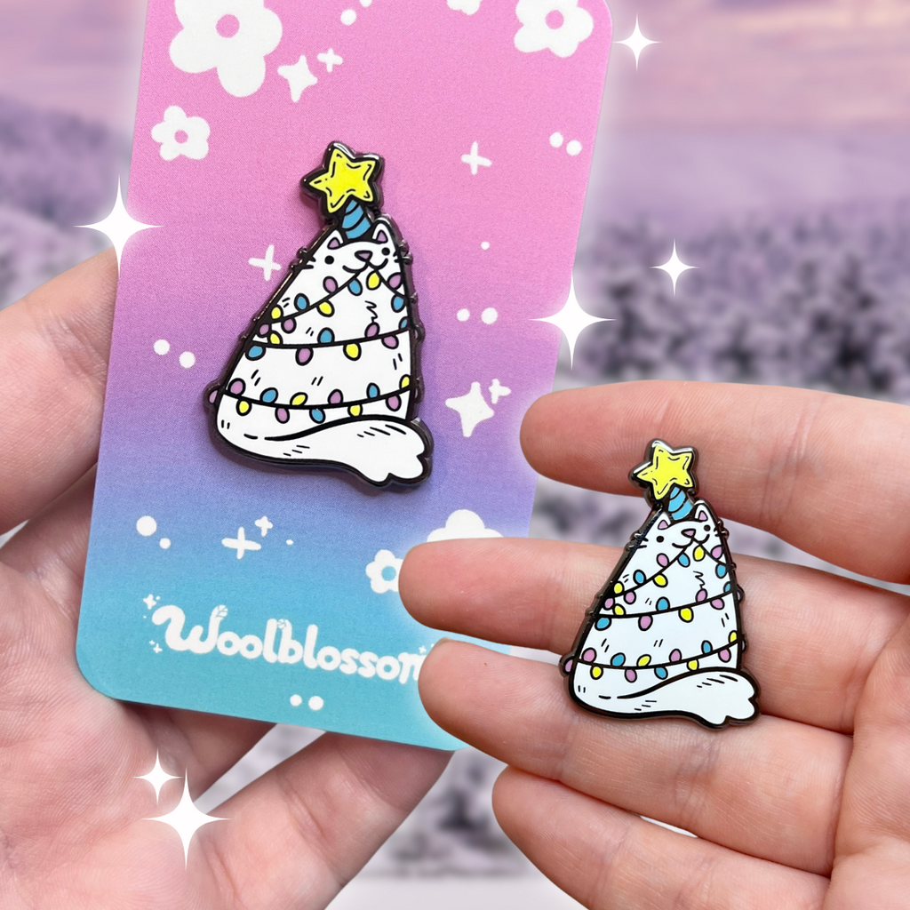 An enamel pin of a white triangular cat, with christmas lights wrapped around them, and a star on their head