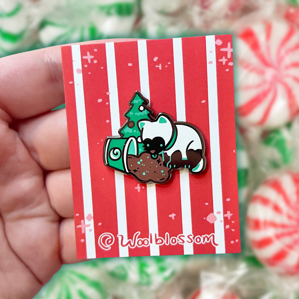 An enamel pin of a white cat with black markings licking spilled hot chocolate