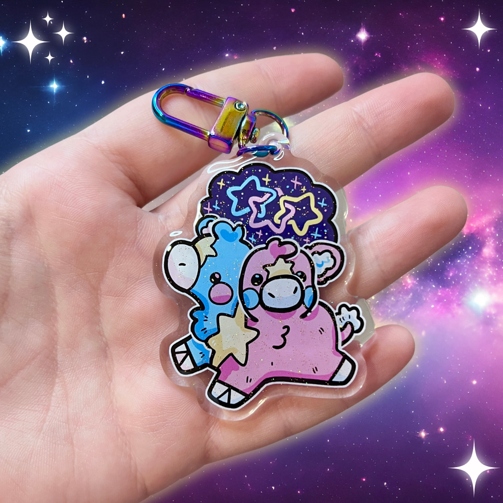 A keychain charm of a two headed calf, one is blue and one is pink, with a yellow star pattern connecting them and on each forehead. On top is a starry sky with three interlocking stars, one blue, one pink, and one yellow.