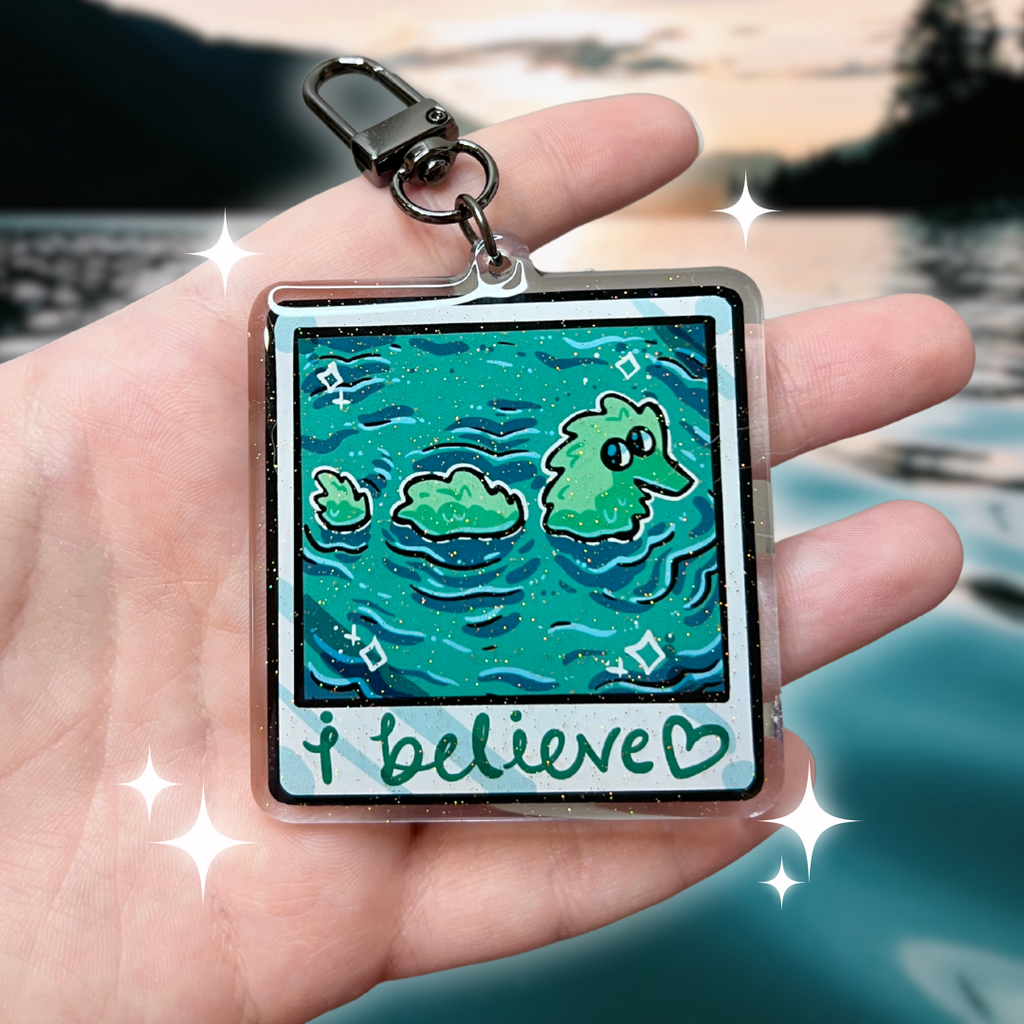 A keychain that looks like a camera photo, hand written text at the bottom that says "i believe <3", in the photo is lake water with a fuzzy worm lochness monster