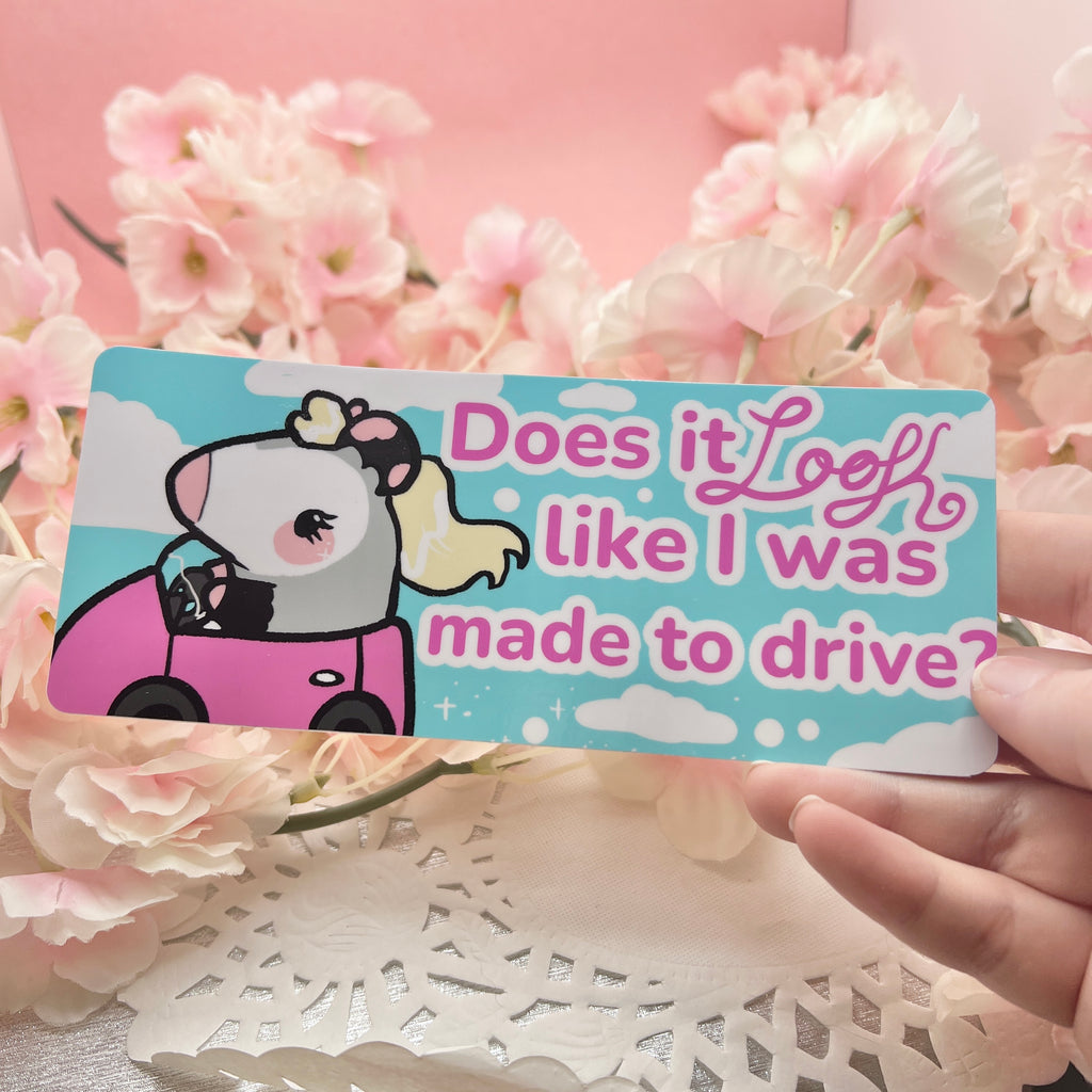 A bumper sticker of a blonde feminine opossum driving with text that says "does it look like I was made to drive"