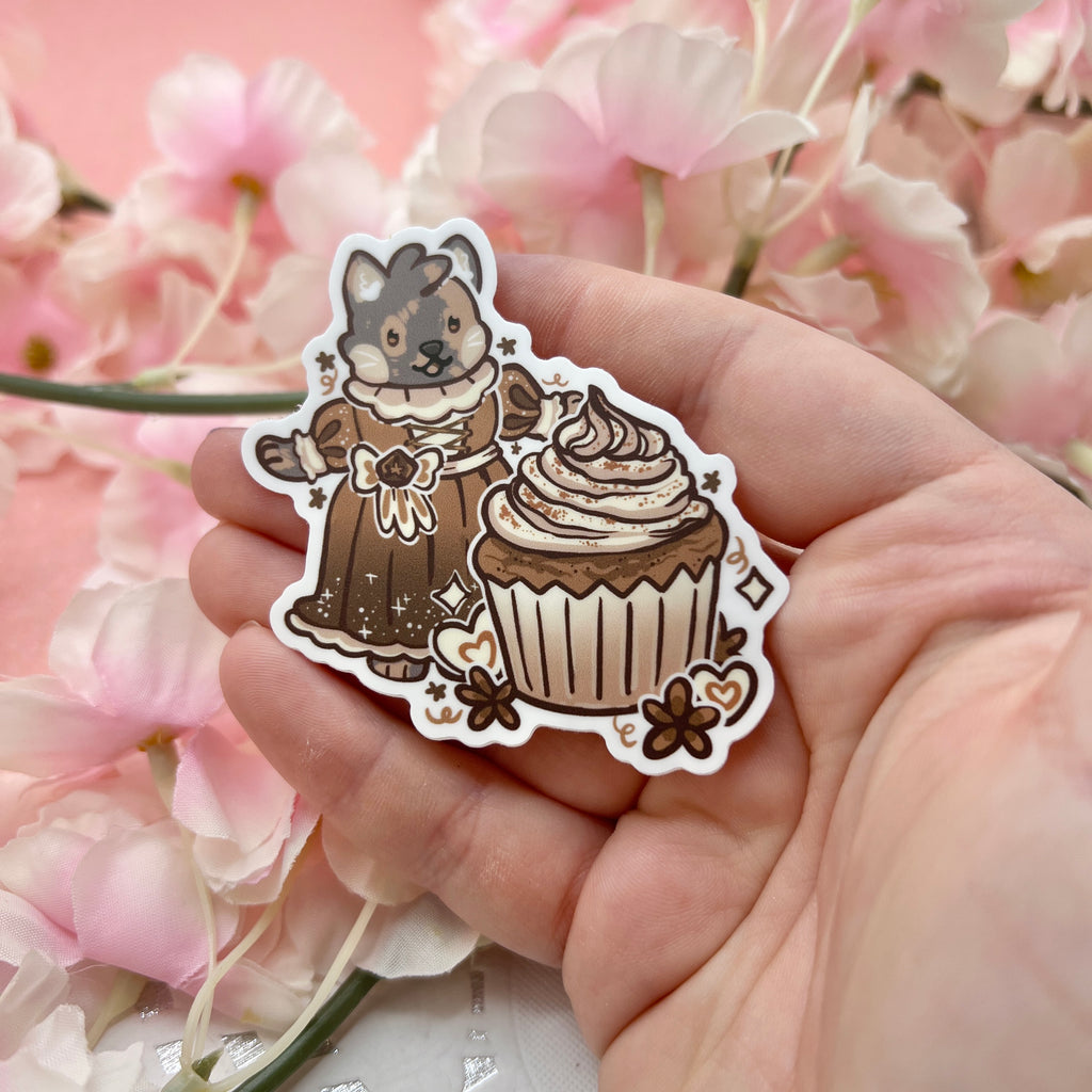 A sticker of my cat Chai dressed in a renaissance/victorian dress, in front of a chai cupcake, and anisse themed