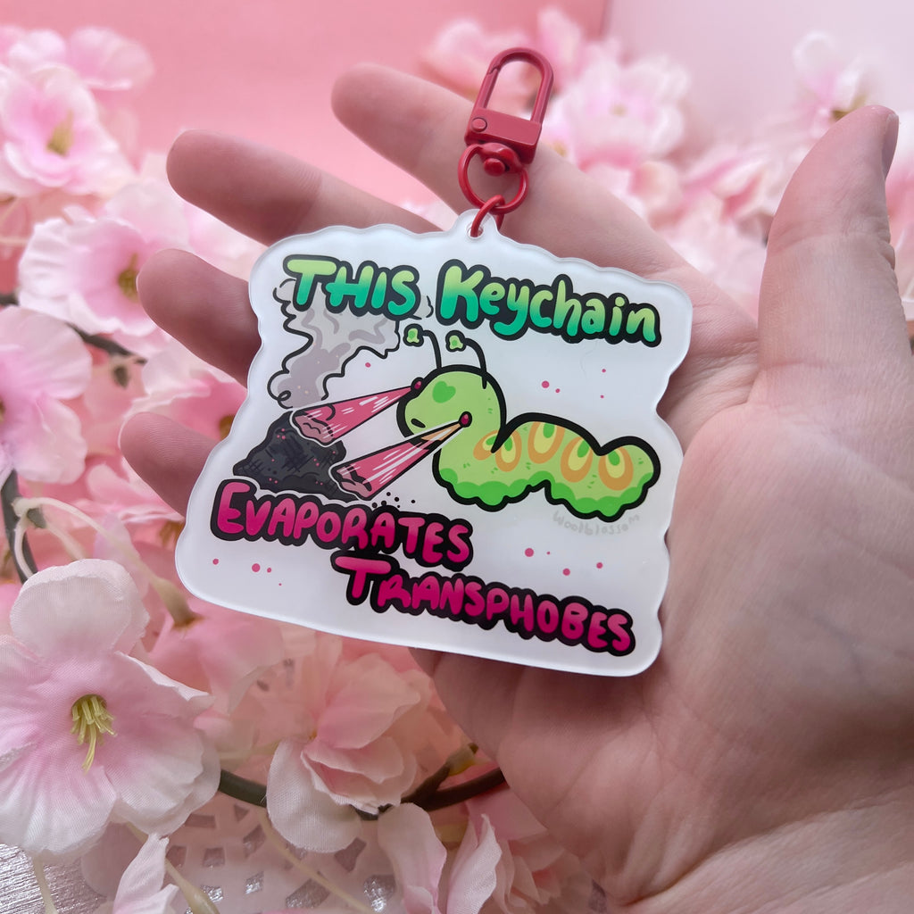 A keychain of a green worm shooting lasers out of its eyes onto a pile of ash with text that says "this keychain evaporates transphobes"