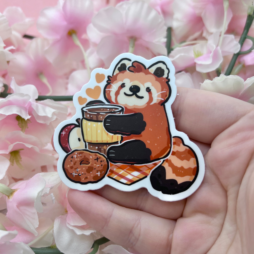 A sticker of a red panda holding a coffee drink with an apple, apple cider doughnut, on a warm toned plaid blanket