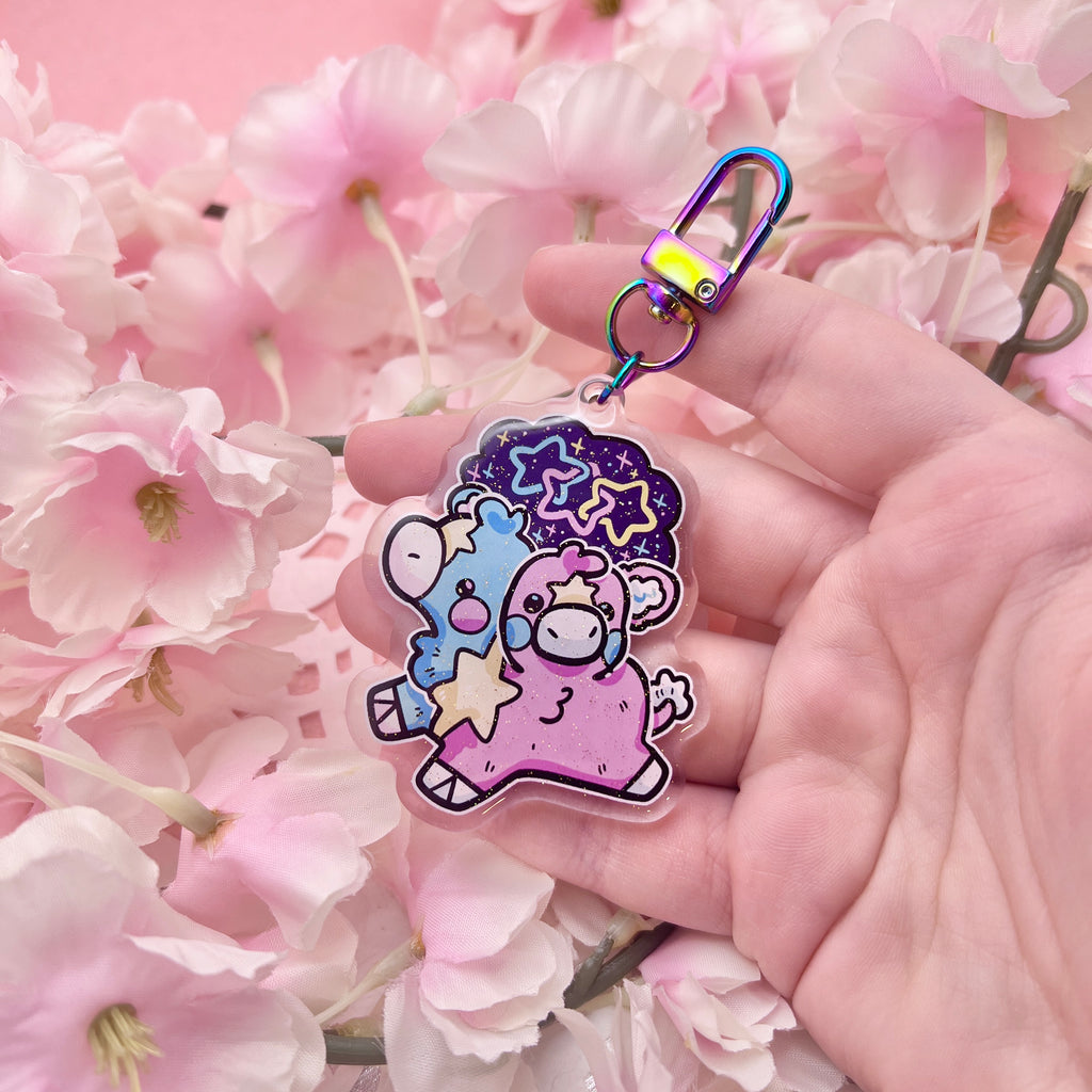 A keychain charm of a two headed calf, one is blue and one is pink, with a yellow star pattern connecting them and on each forehead. On top is a starry sky with three interlocking stars, one blue, one pink, and one yellow.