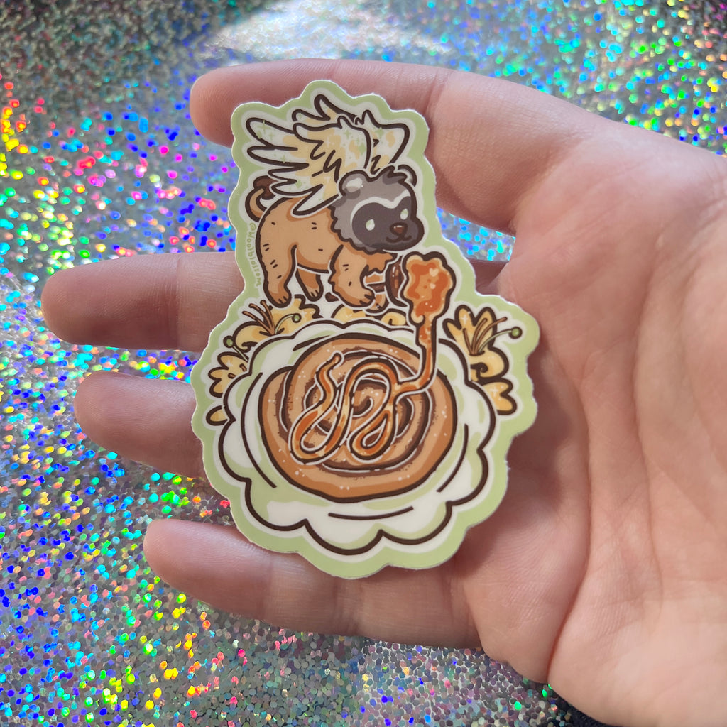 Hand holding a sticker of a badger gryffin pouring honey over a cinnamon roll with honeysuckles, against a glittery background.