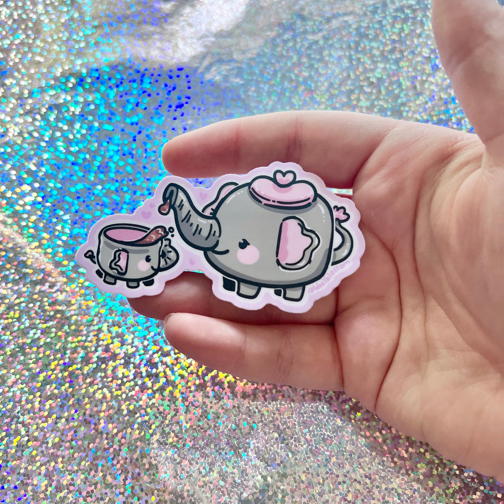 Hand holdinga sticker of a teapot elephant and her baby teacup elephant on a glittery background