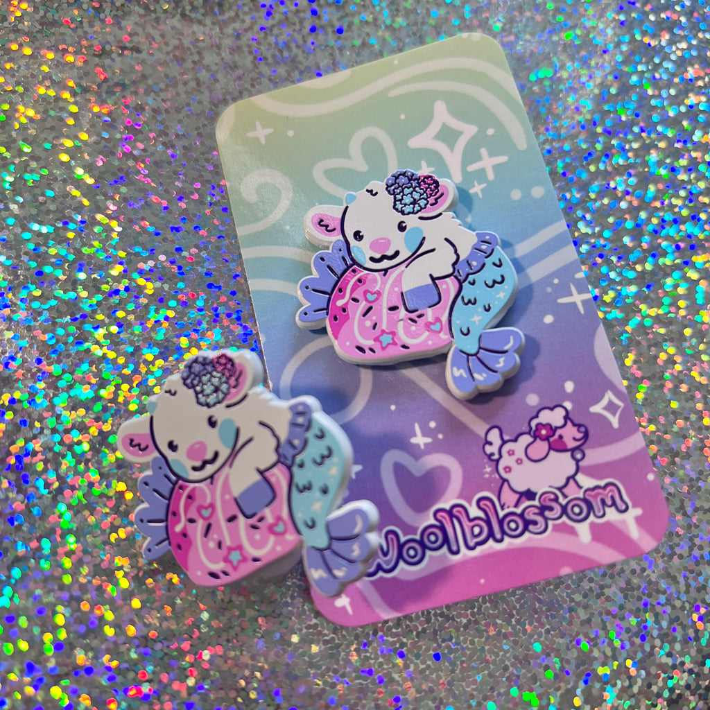 Enamel pin of a mer-goat holding a strawberry on a glittery background with 'Woolblossom' branding.