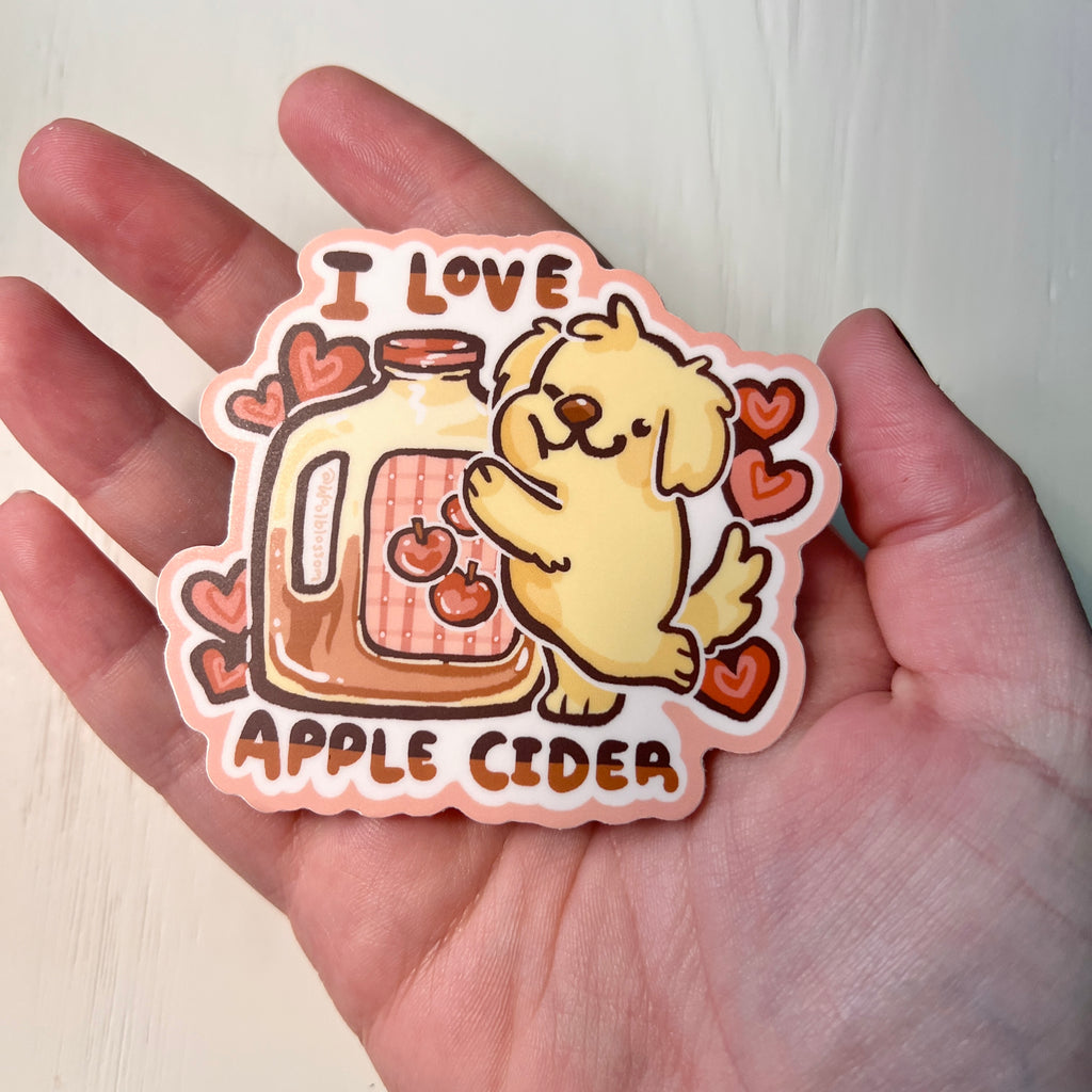 Sticker with a dog and apple cider design held in a hand against a white background