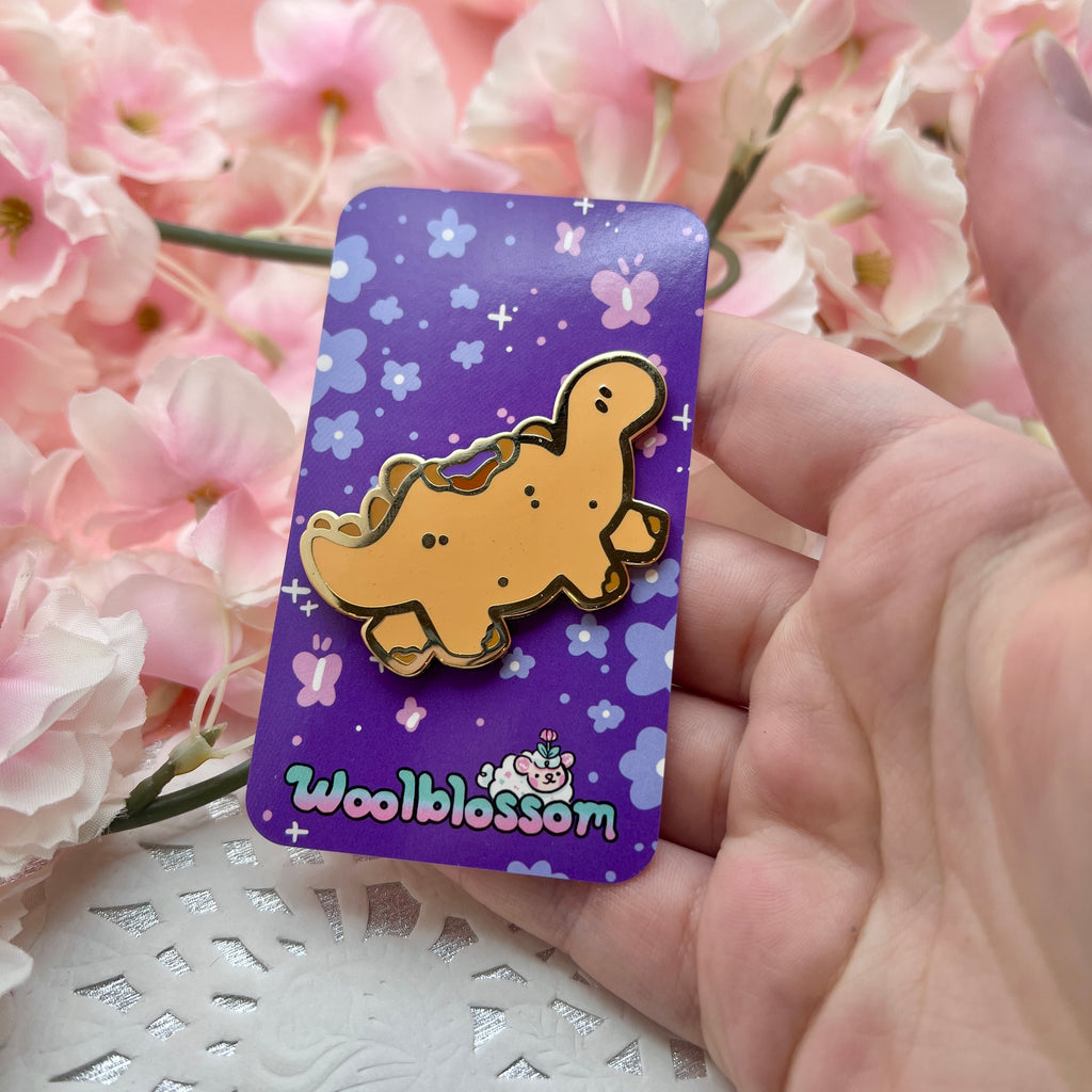 A gold enamel pin of a tan colored stegosaurus inspired dino that resembles a PBJ sandwich, the crustless kind, with a bite taken out of its back so you see the PB and jelly