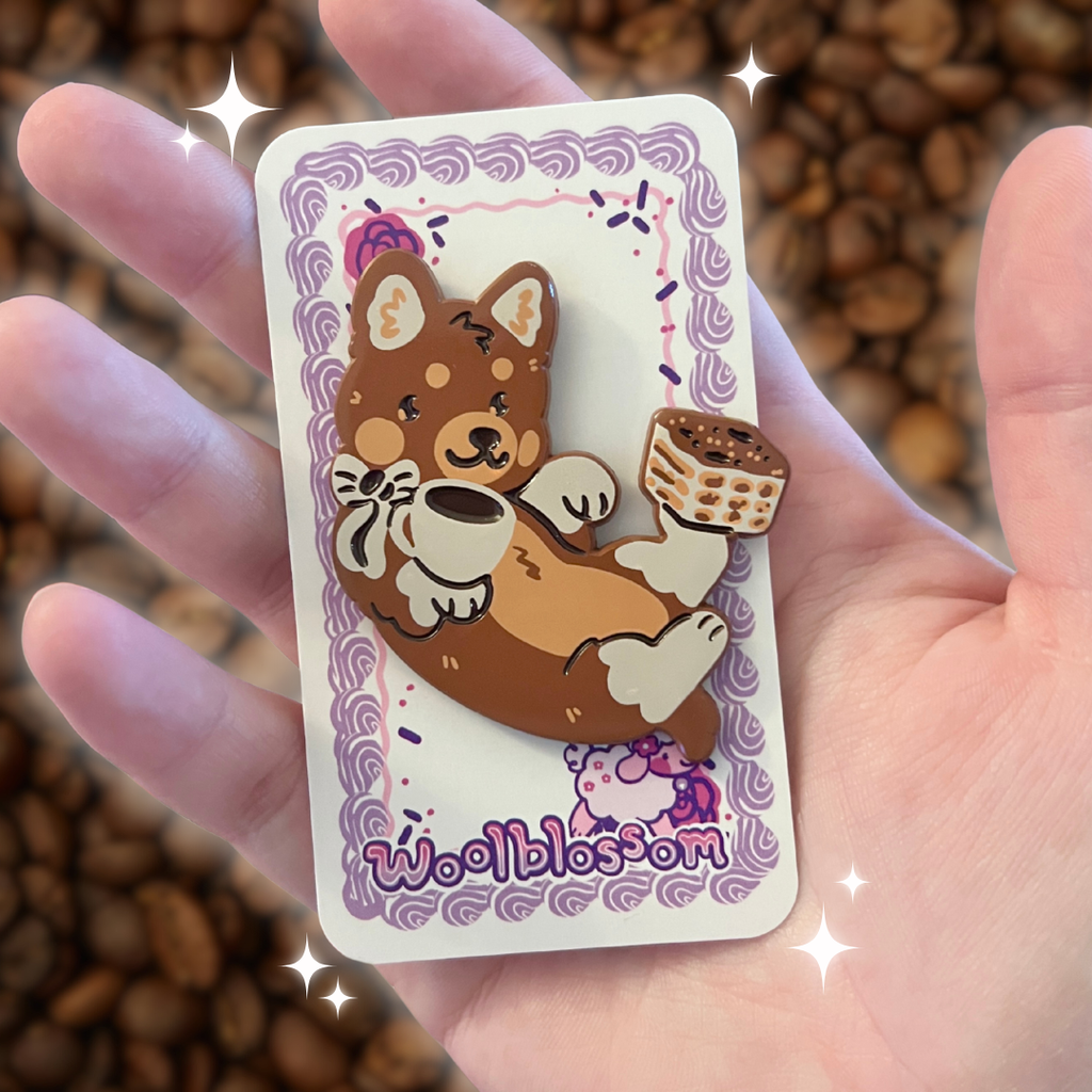 An enamel pin of a brown dog with a tiramisu and espresso on his paws