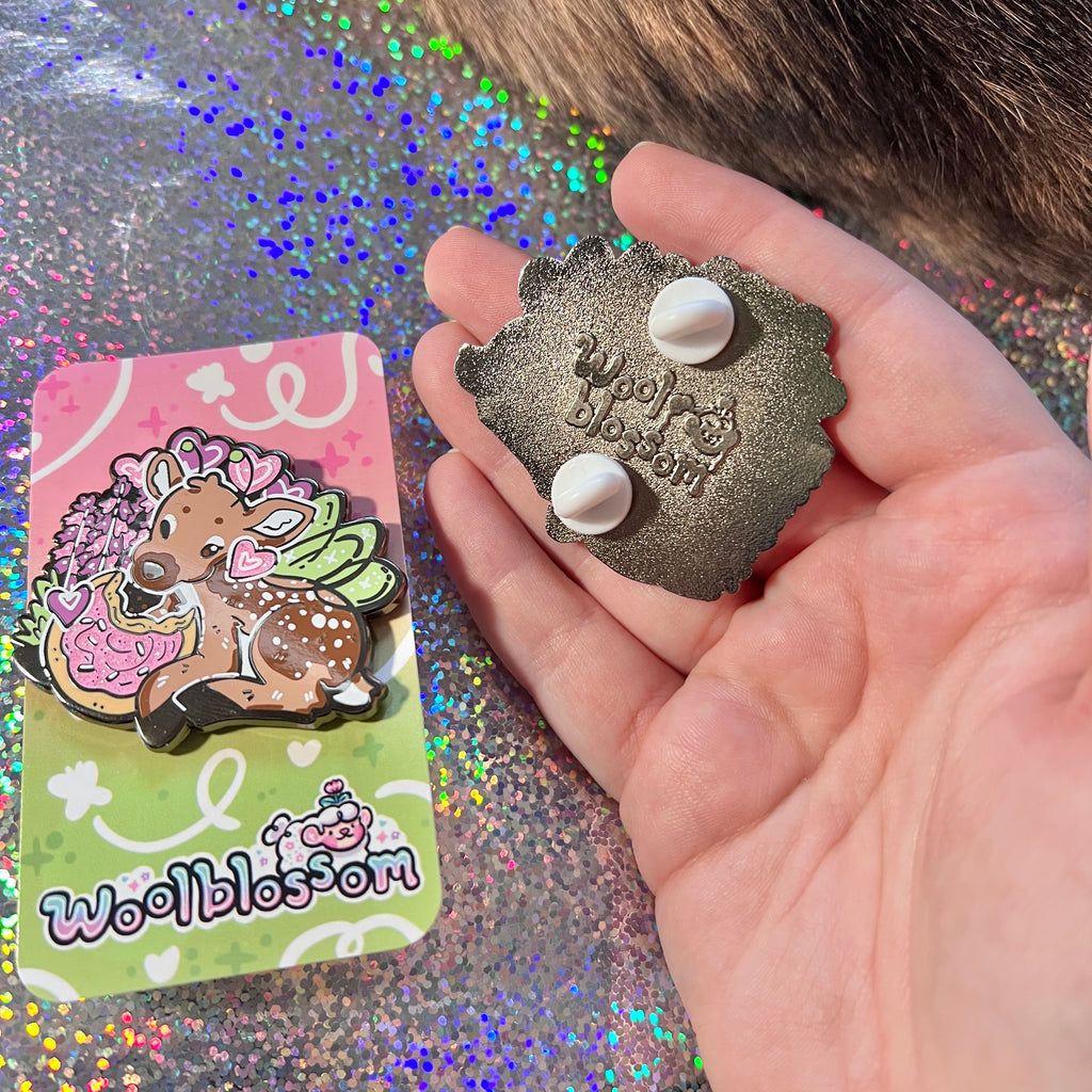 Hand holding a metallic pin with 'woolblossom' branding, next to its packaging on a glittery background.