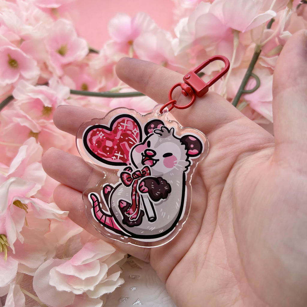 a keychain of an opossum happily holding a red heart lollipop