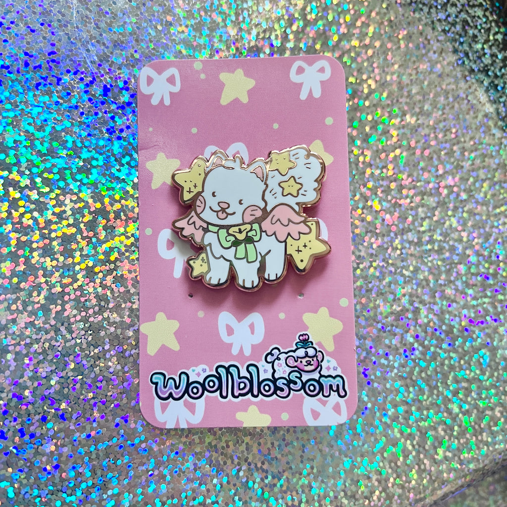 An enamel pin of a white cat with pink wings and yellow stars on a pink card with 'woolblossom' branding against a glittery background.