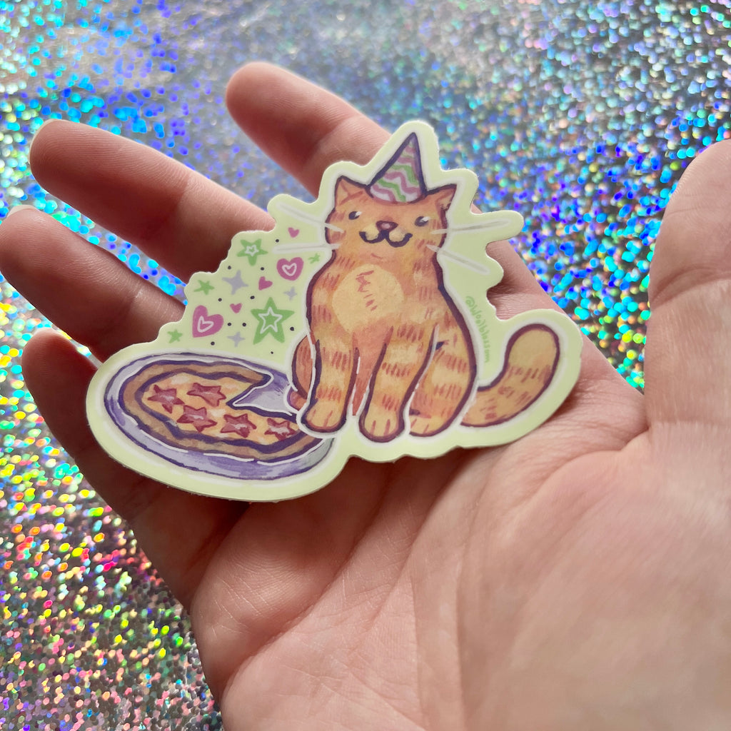 Hand holding a sticker of a cat with a party hat against a glittery background