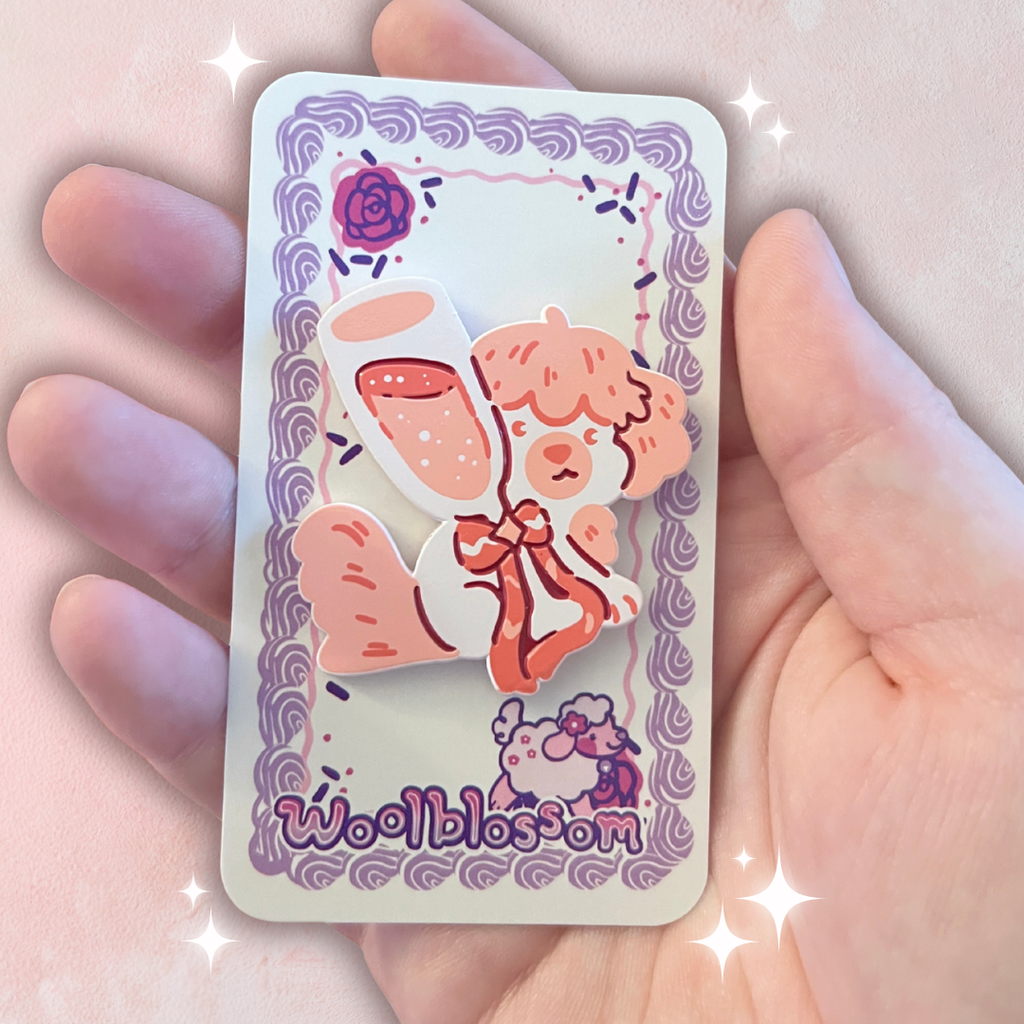 An enamel pin of a pale pink dog behind a glass of rose